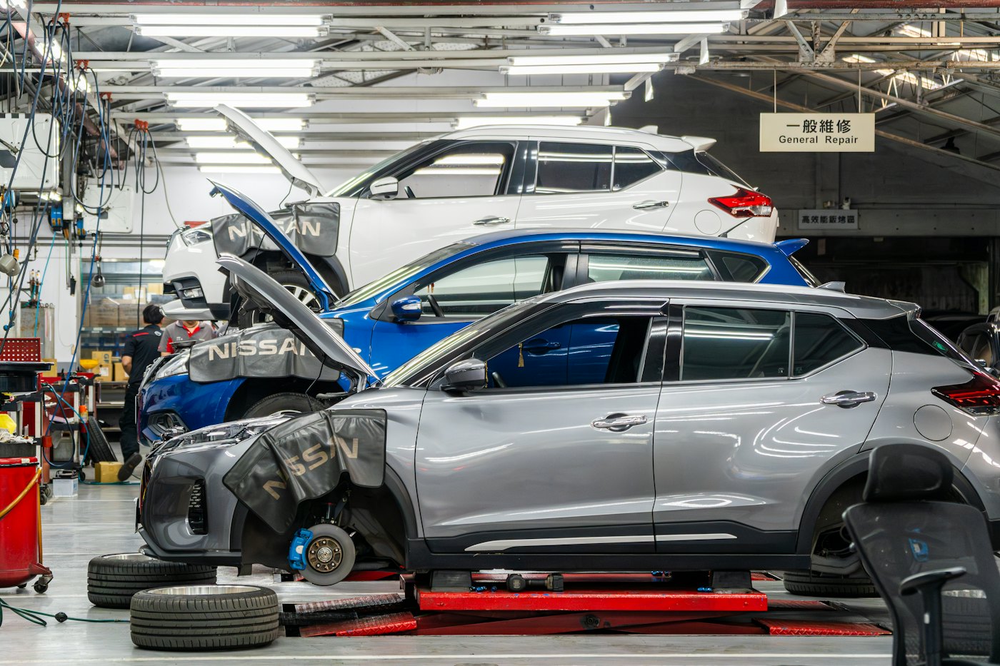Two cars side by side on low lifts in a bright workshop, one with its front wheel off.