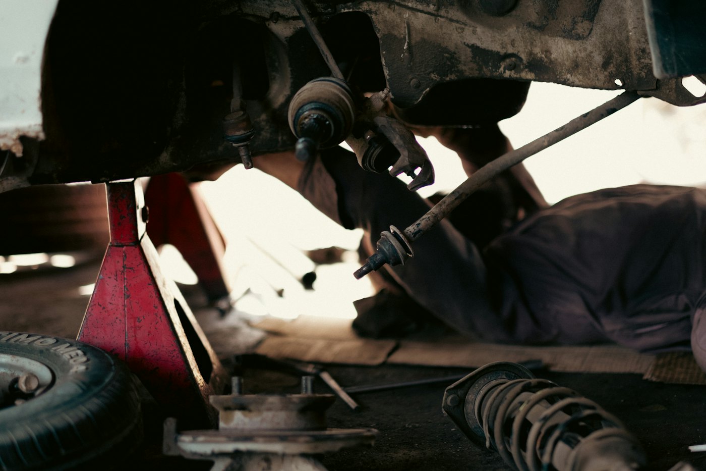 Under a car on stands: a mechanic working behind the front suspension, with a removed coil spring on the floor.