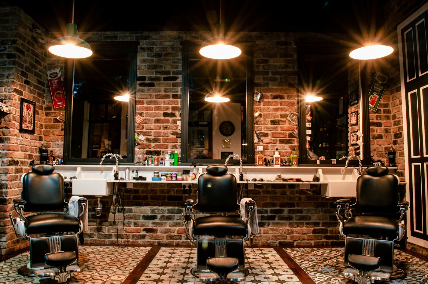 Three black leather barber chairs facing mirrors on a brick wall, lit by warm pendant lamps over a patterned tile floor