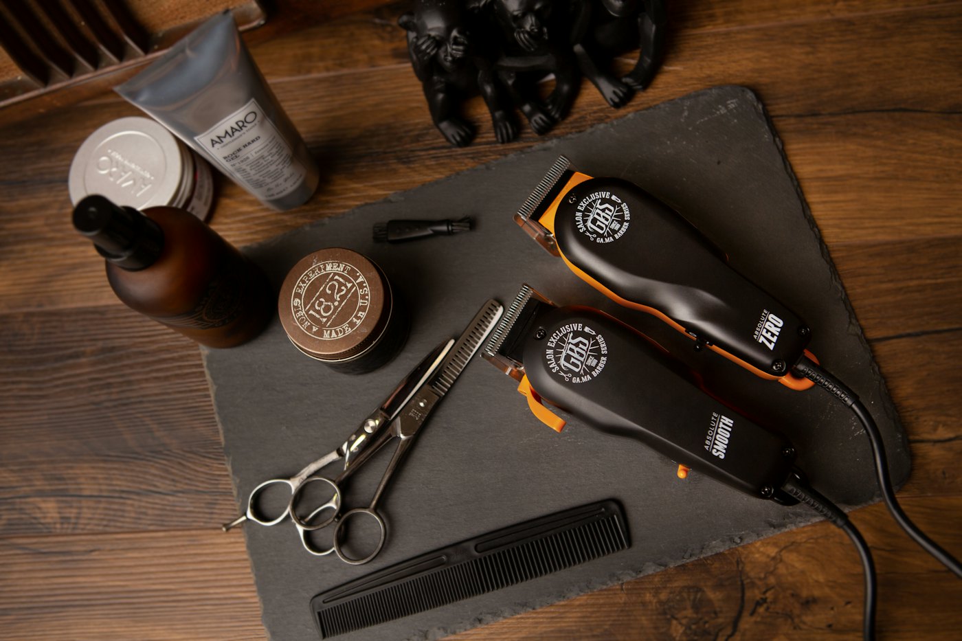 Two clippers, scissors, a comb and jars of pomade laid out on a dark slate mat on a wooden counter