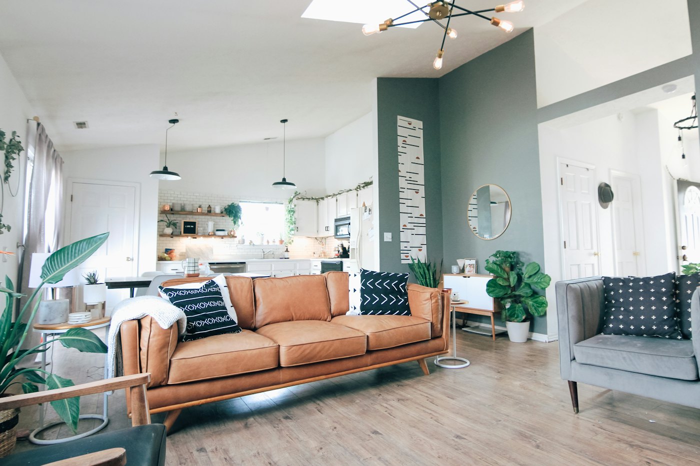 A bright, tidy open-plan living room with a tan leather sofa, house plants and a kitchen behind it