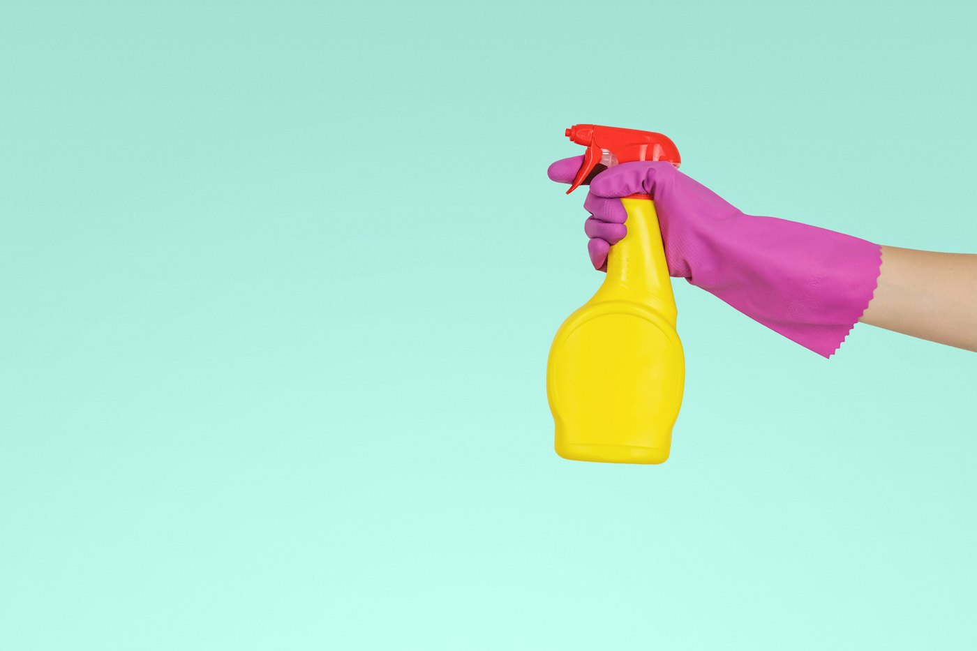 A hand in a pink rubber glove holding a yellow spray bottle against a mint green wall