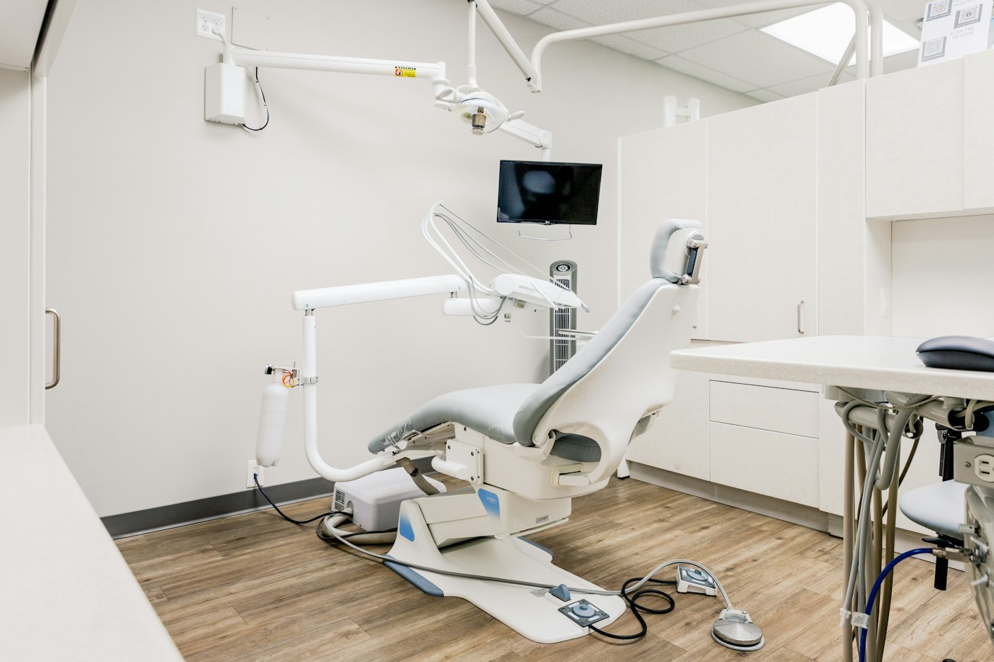 A bright treatment room with a white dental chair, an overhead light on a long arm, pale cabinets and a wood floor.