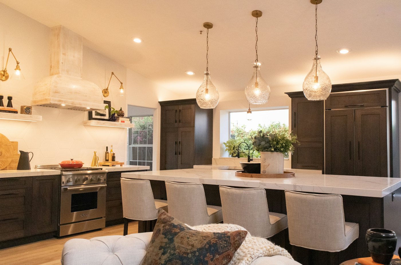 A kitchen in the evening with three glass pendant lights glowing over a white stone island, dark cabinets behind.
