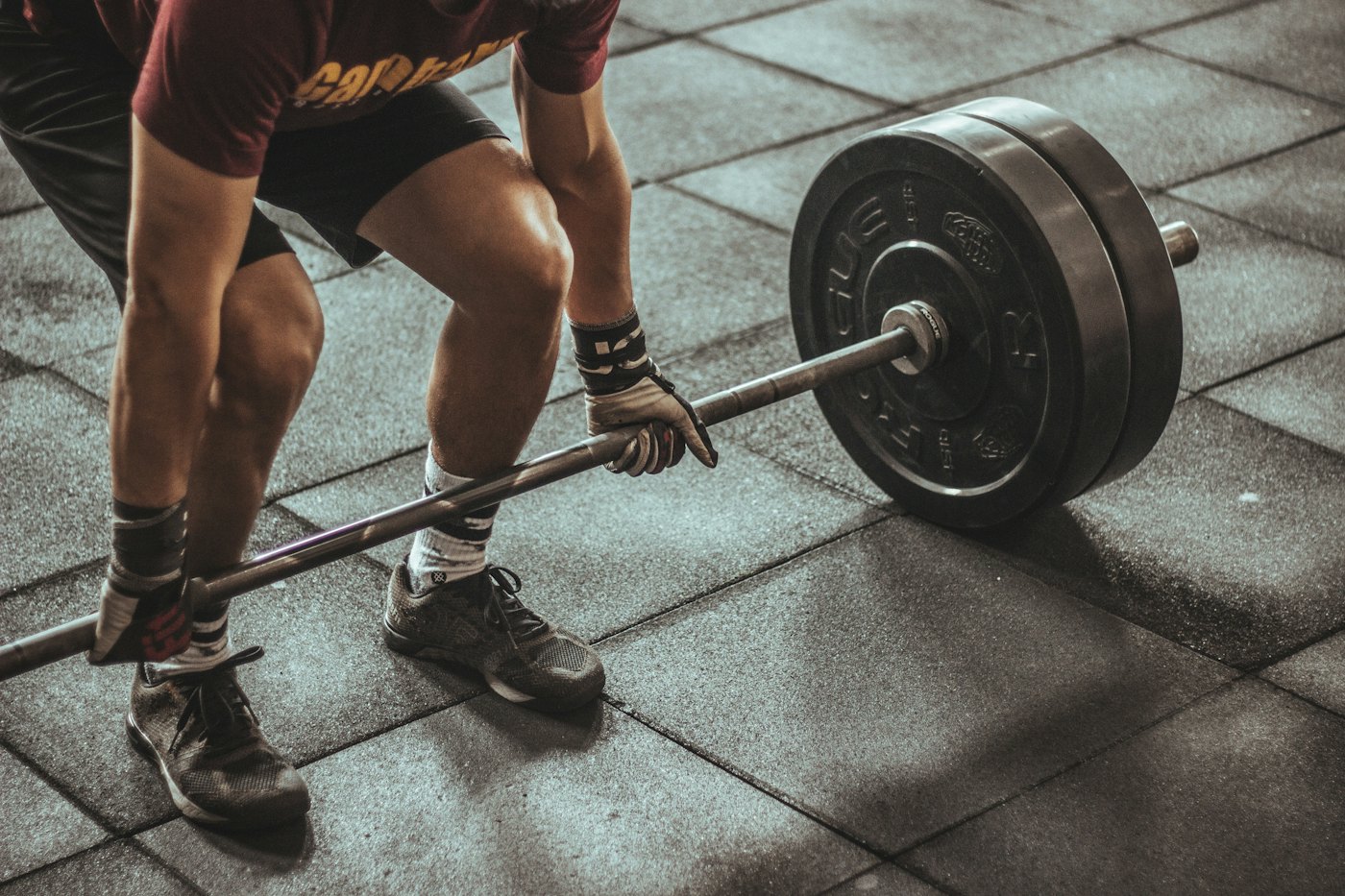 A lifter in gloves and wrist wraps grips a barbell loaded with black bumper plates, about to pull it from a rubber tile floor.