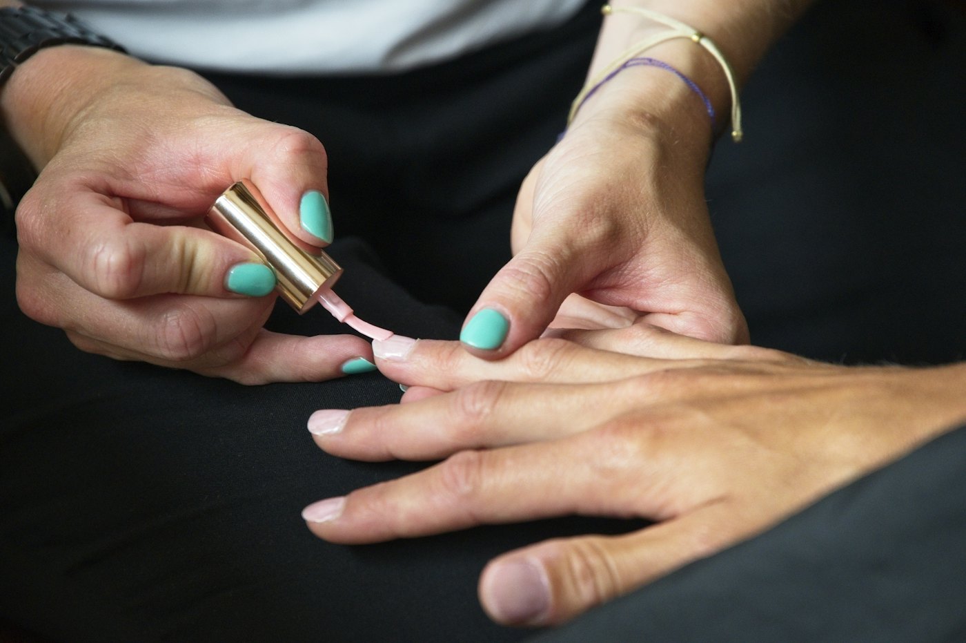 A nail technician with teal nails brushing pale pink polish onto a client's fingernail