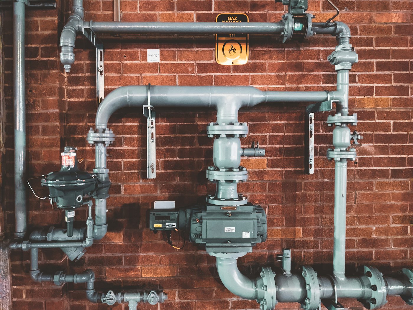 Grey steel pipes, valves and a meter fixed to a red brick wall