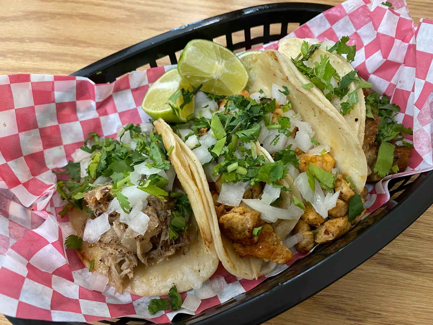 Three tacos on corn tortillas with cilantro, white onion and lime wedges in a basket lined with red and white checkered paper