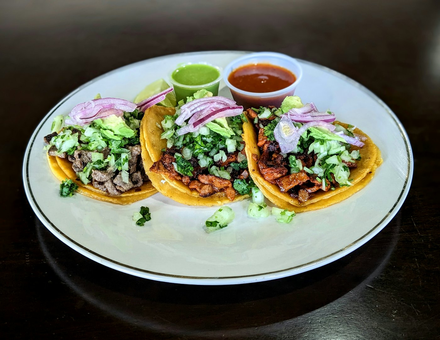Three tacos with avocado and pickled red onion on a white plate, with cups of green and red salsa